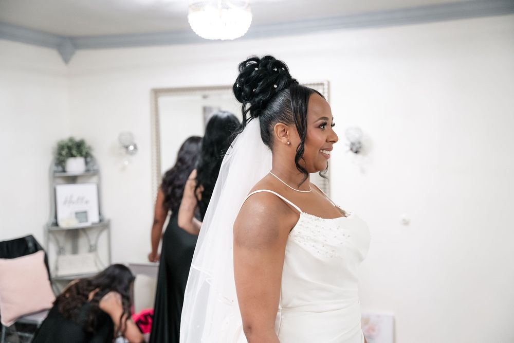 A bride in an elegant white wedding dress and veil smiles while getting ready in a bright room before her ceremony.