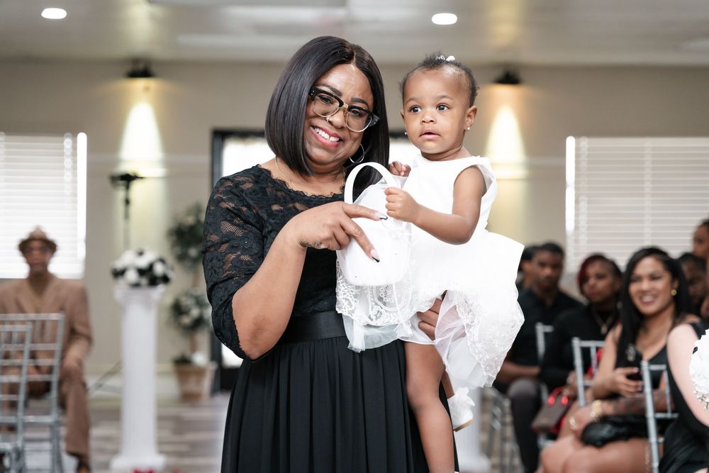 A celebratory indoor gathering shows a heartwarming moment between an adult wearing a dark dress and a toddler in white.