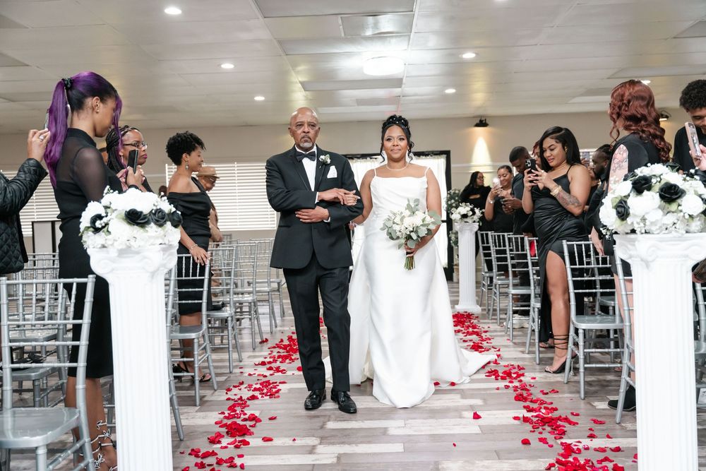 A wedding ceremony aisle decorated with red rose petals and white chiavari chairs in an elegant indoor venue.