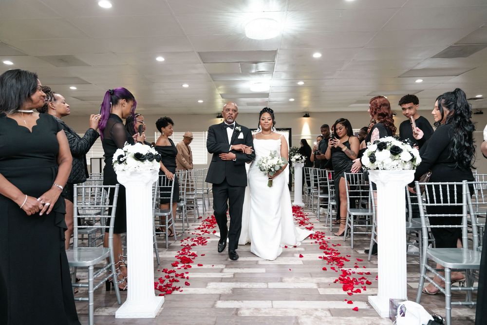 Wedding ceremony scene with decorated aisle lined with white pillars and rose petals in an indoor venue.