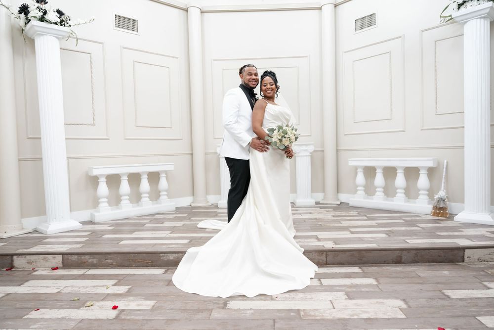 A couple stands together in white wedding attire against a classical white stone balustrade setting.