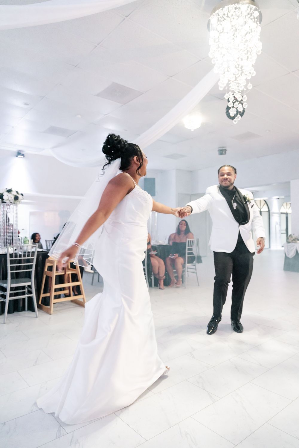 A couple shares their first dance at their wedding reception in an elegant white room with a crystal chandelier above.
