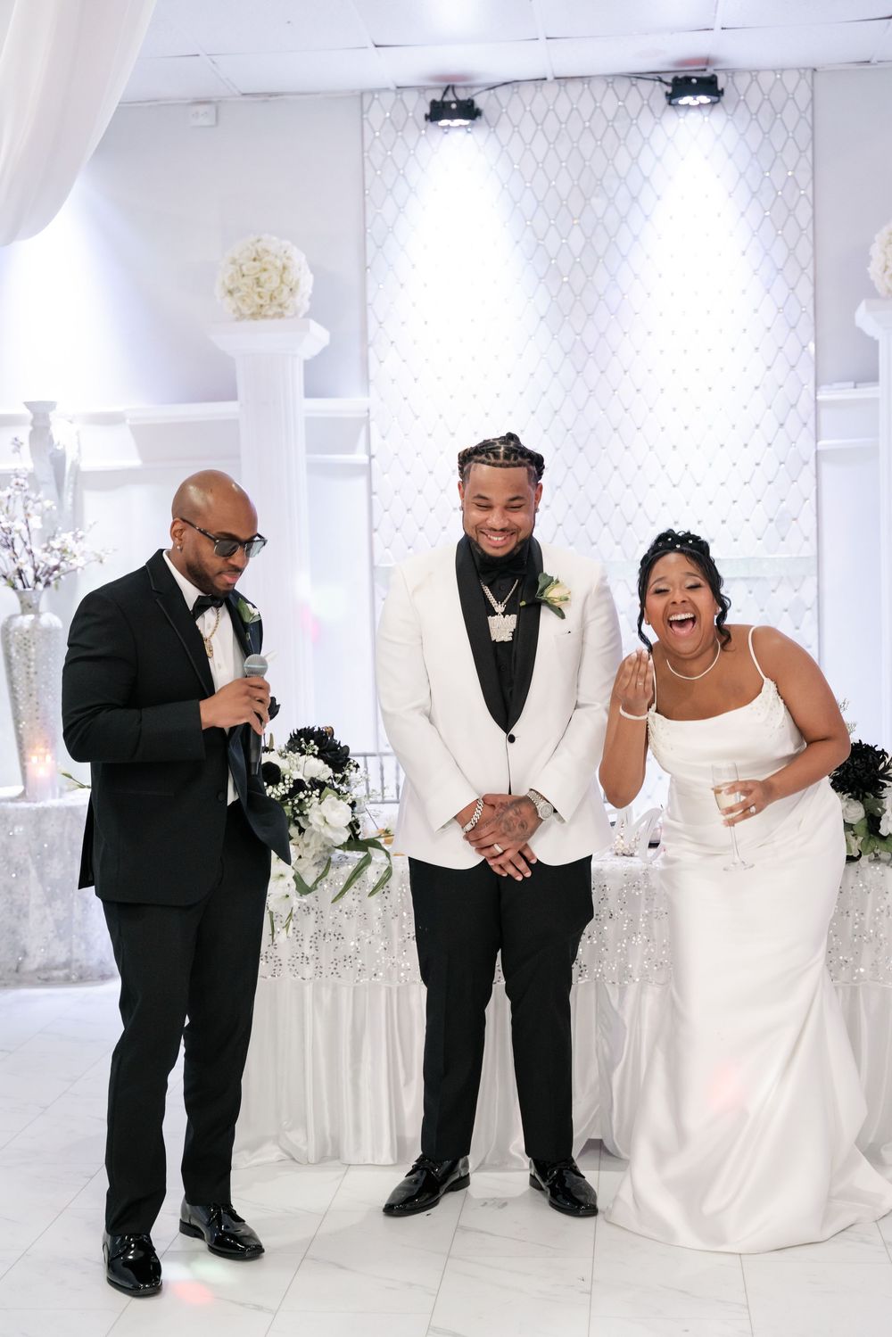 Three people in formal wedding attire share a joyful moment together at an elegant white reception venue.
