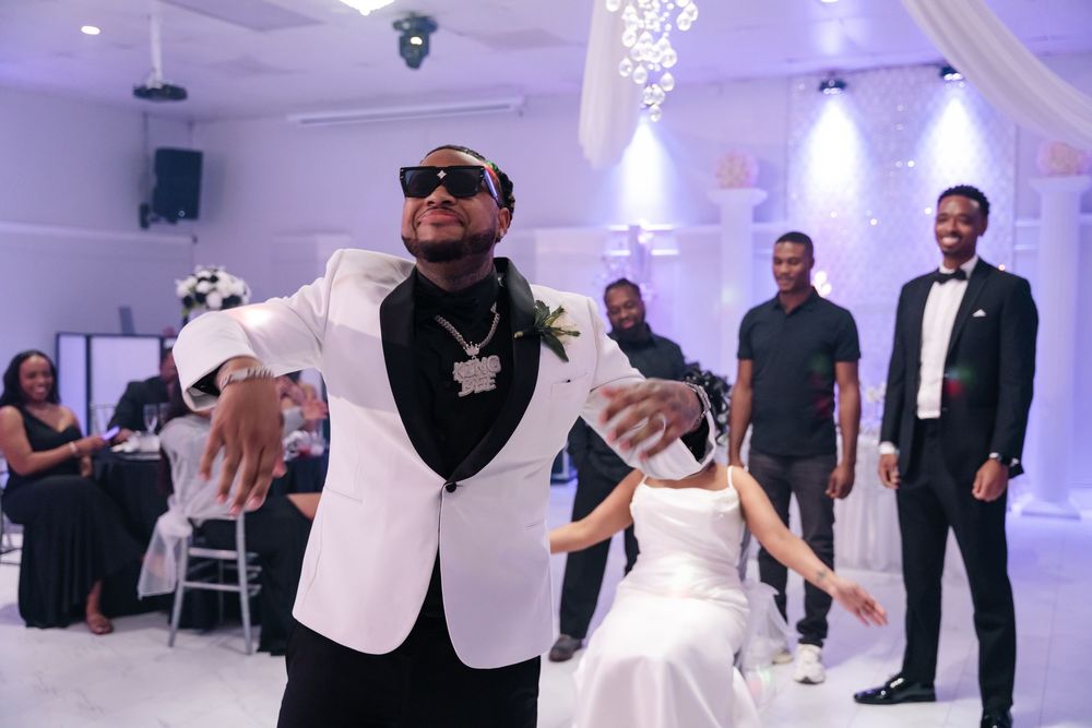 People dance and celebrate at a wedding reception in an elegant white ballroom with purple lighting.