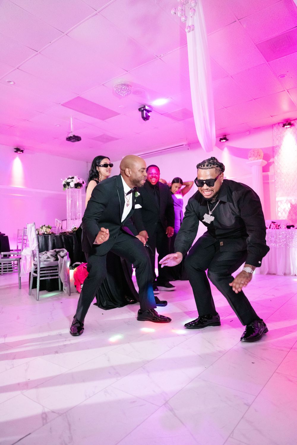 Groomsmen in black suits striking dramatic poses while crawling on white floor with pink uplighting at wedding reception.