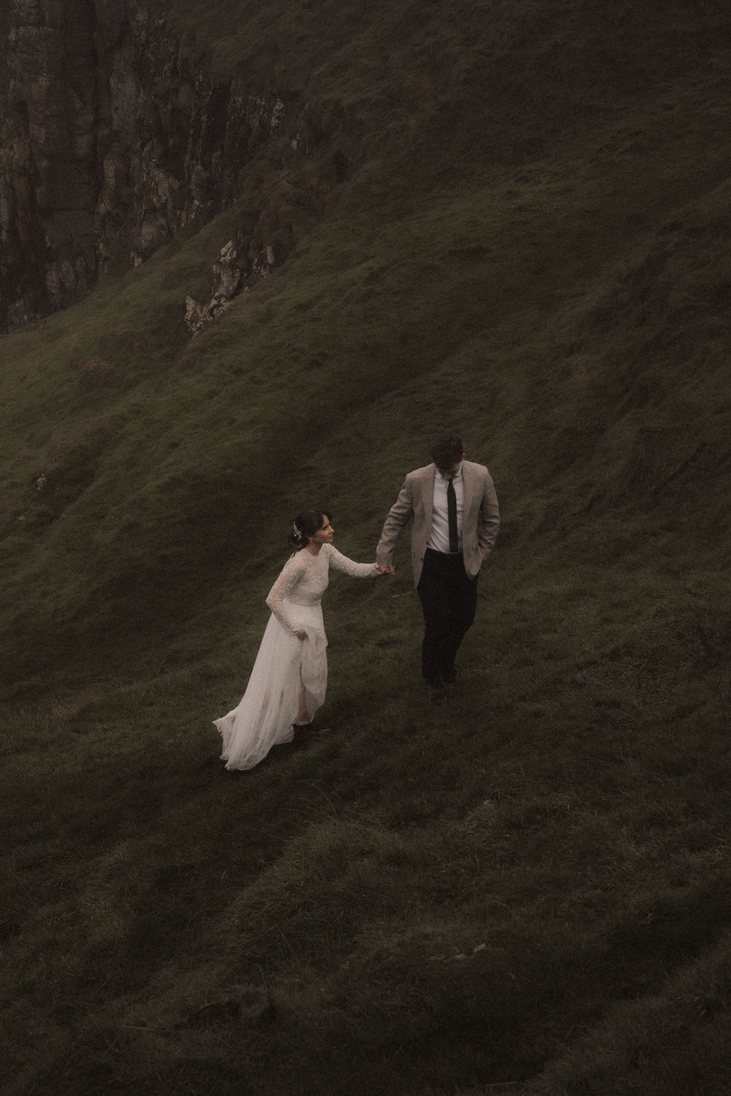 A couple in wedding attire walking together on a misty green hillside slope at dusk.