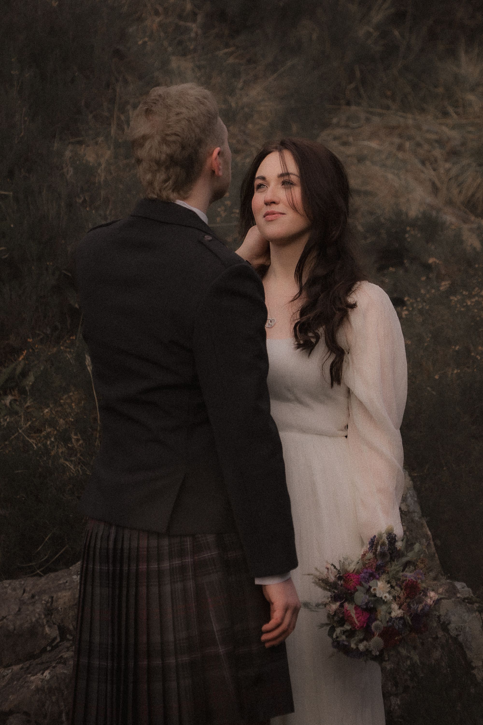 A couple shares an intimate wedding moment against a moody backdrop, with the groom in traditional Scottish kilt attire.