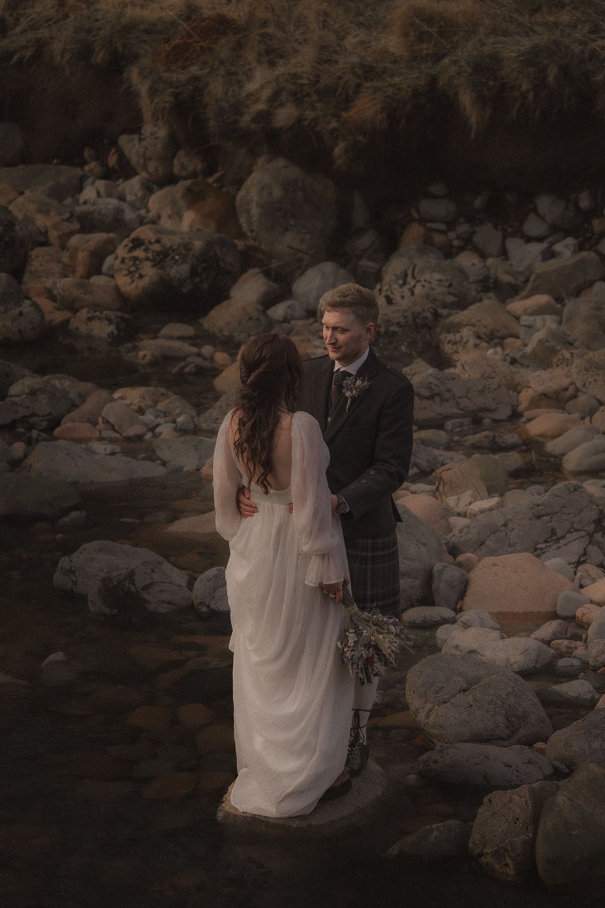 A couple in wedding attire shares intimate moments among rocky terrain at sunset in this moody photoshoot sequence.