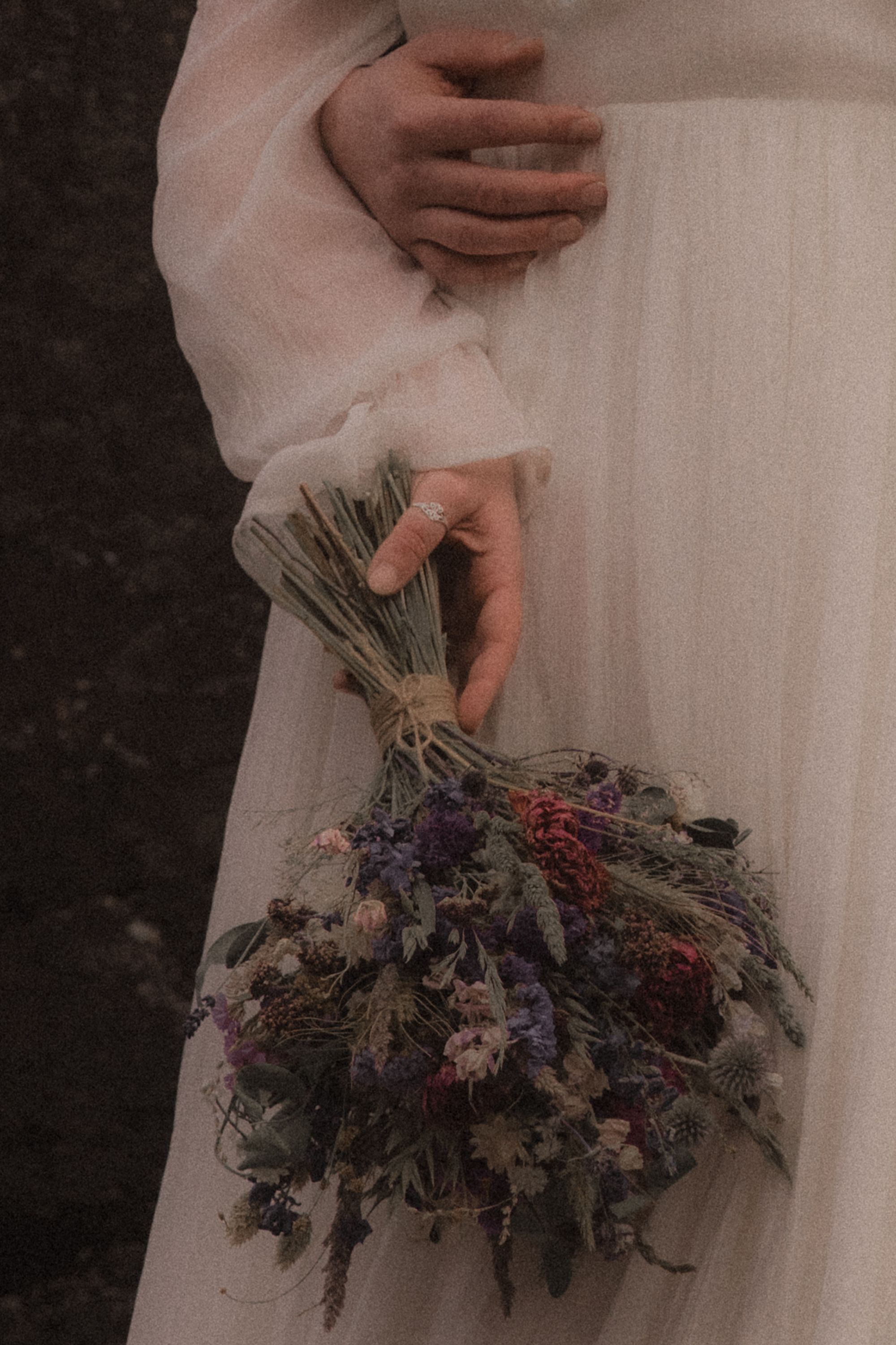 A person in white dress holds a dried flower bouquet featuring purple and red blooms against a dark background.