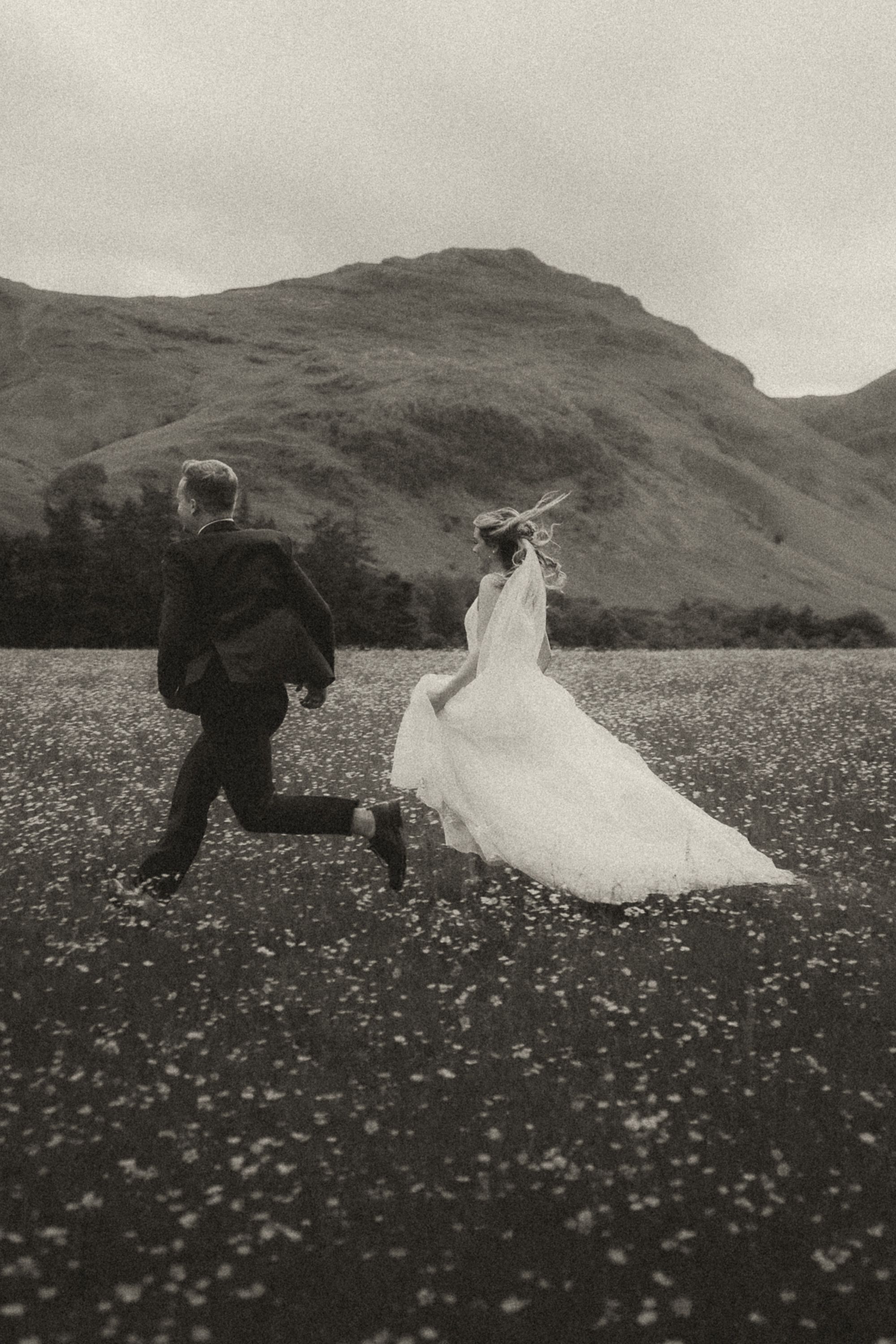 A couple in wedding attire runs through a picturesque meadow filled with wildflowers against a backdrop of rolling hills.