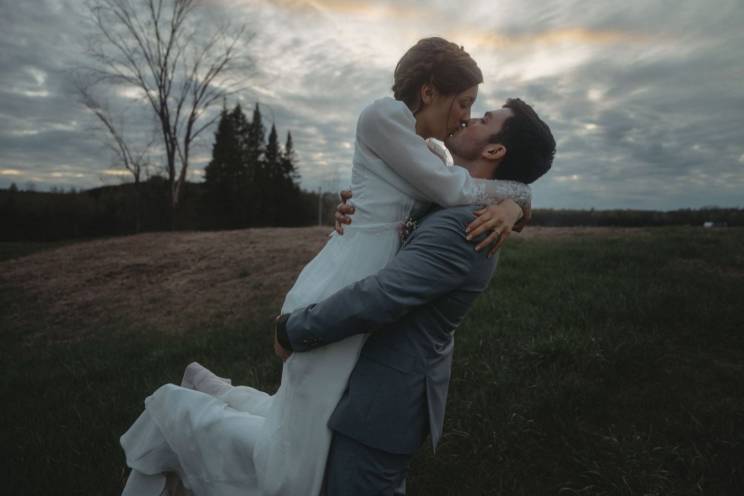 A romantic sunset embrace between newlyweds in a grassy field with trees in the background.