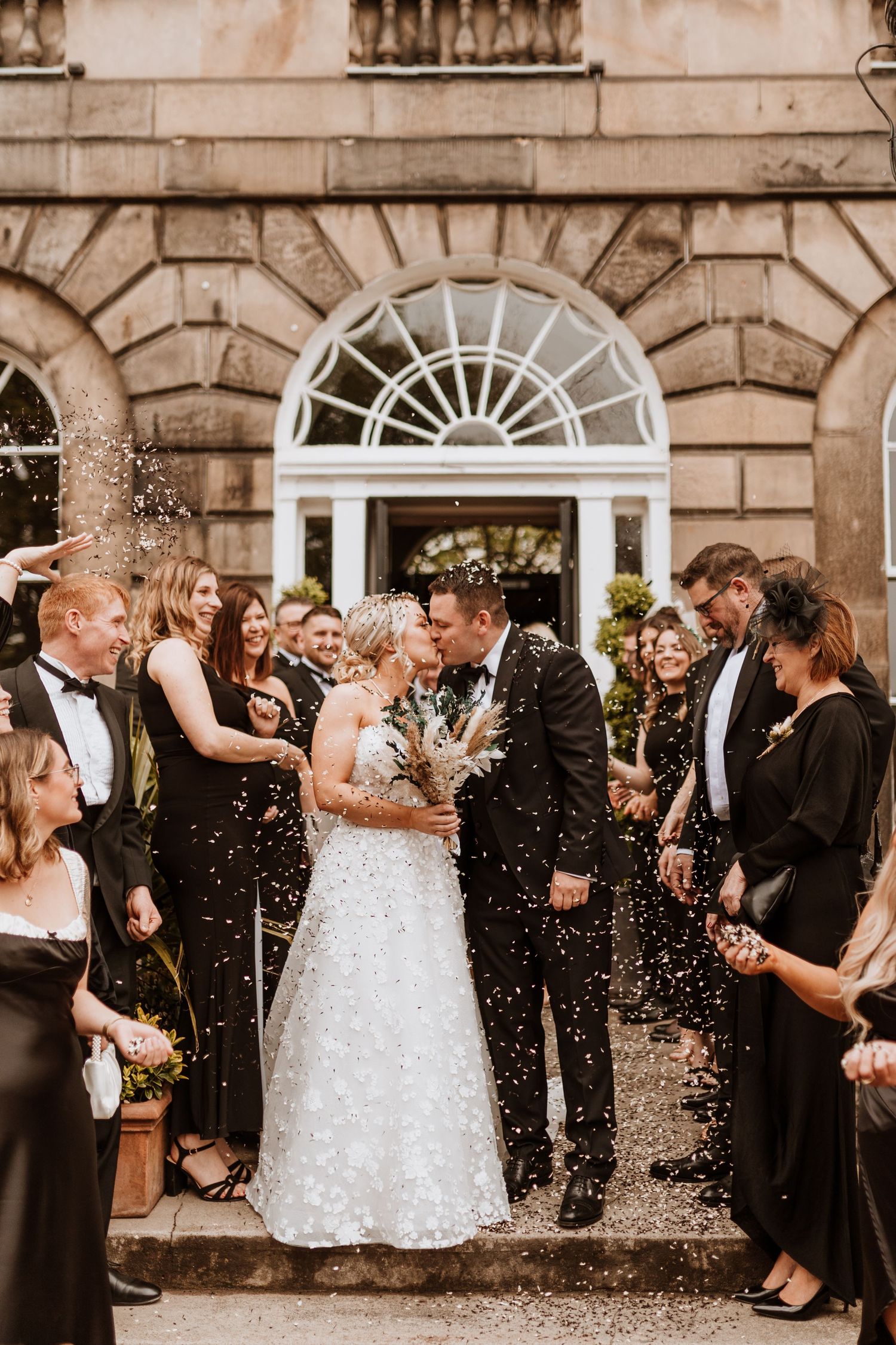 Wedding celebration outside historic building with stone facade as guests shower newlyweds with confetti.