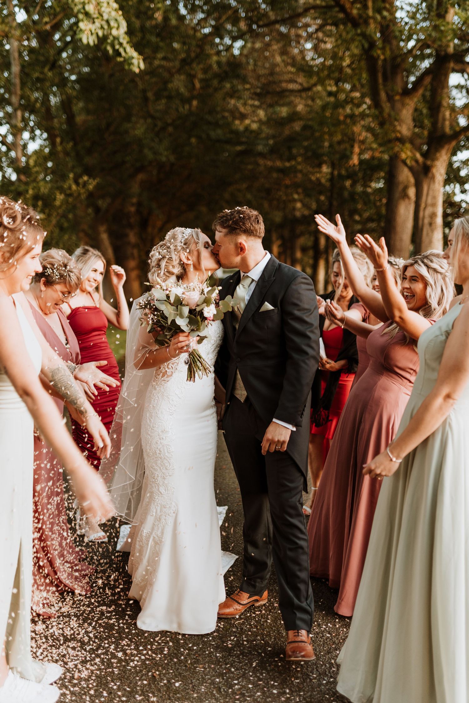 Wedding party celebrates on a tree-lined path as couple shares a tender moment amid joyful bridesmaids in mix of colorful dresses.