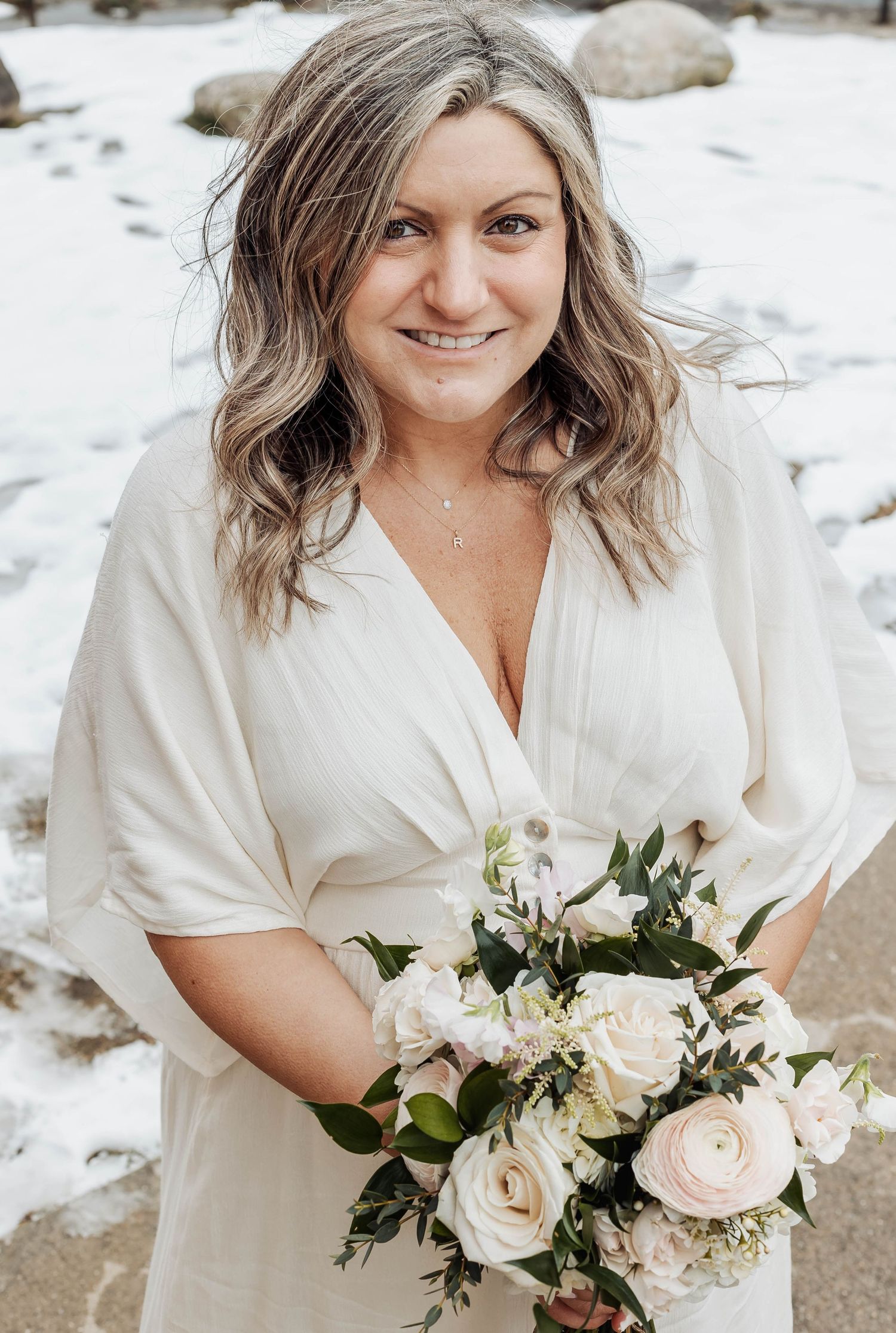 A bride in a white wrap dress holds a romantic bouquet of blush and white roses against a snowy winter backdrop.