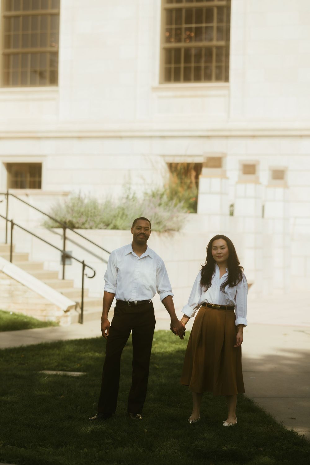 Stylish couple in white shirts and brown pants standing on grass outside a building.