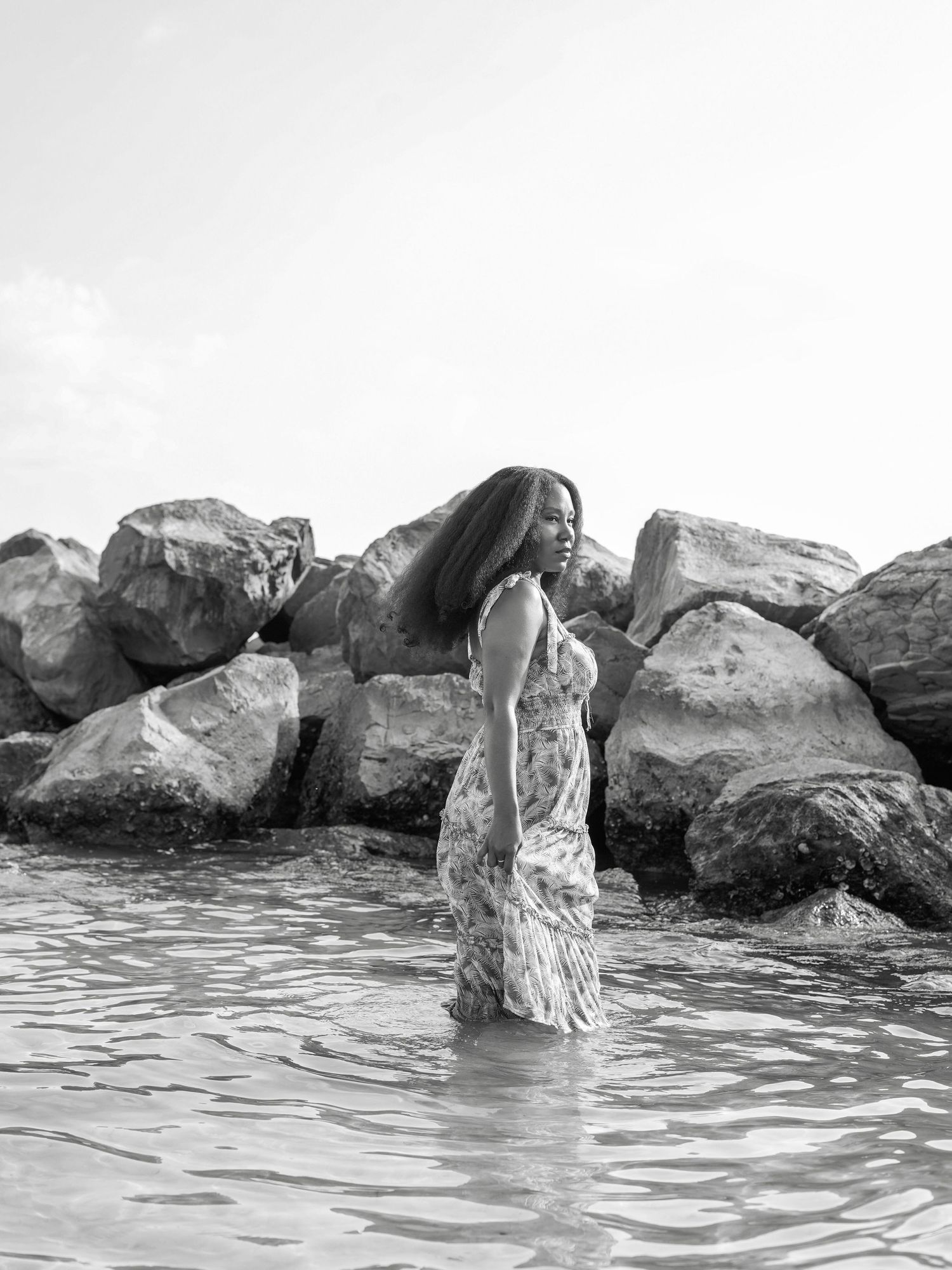 A person in a flowy dress stands in shallow water near large rocks in black and white.