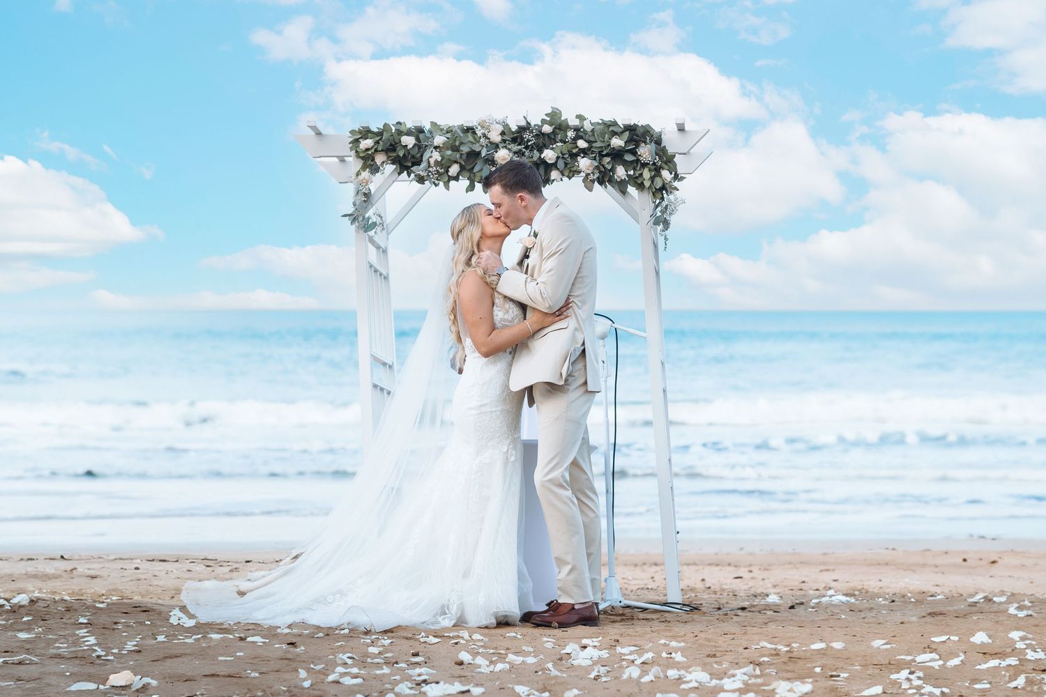 Romantic beach wedding ceremony under floral arch with waves and blue sky in background.