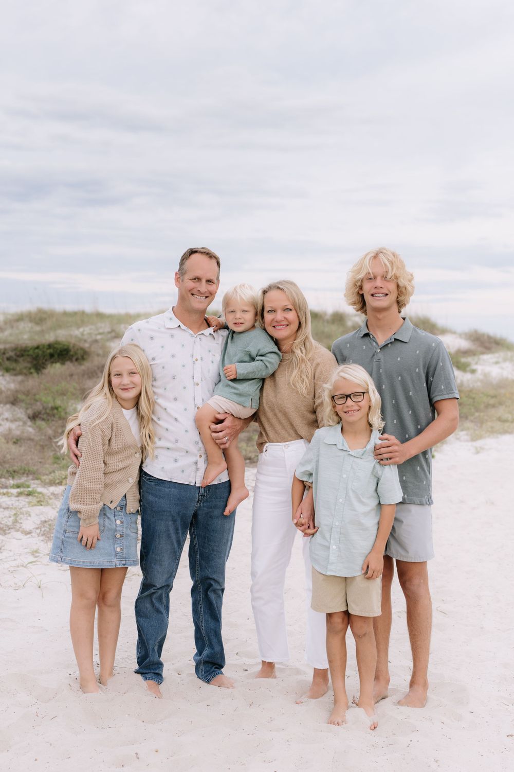 Family stands together on sandy beach with overcast sky in background wearing coordinated beige and white outfits.