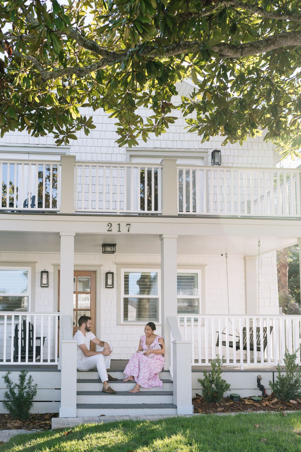 Two people relax on the front porch steps of a classic white house with columns and railing during a sunny afternoon.