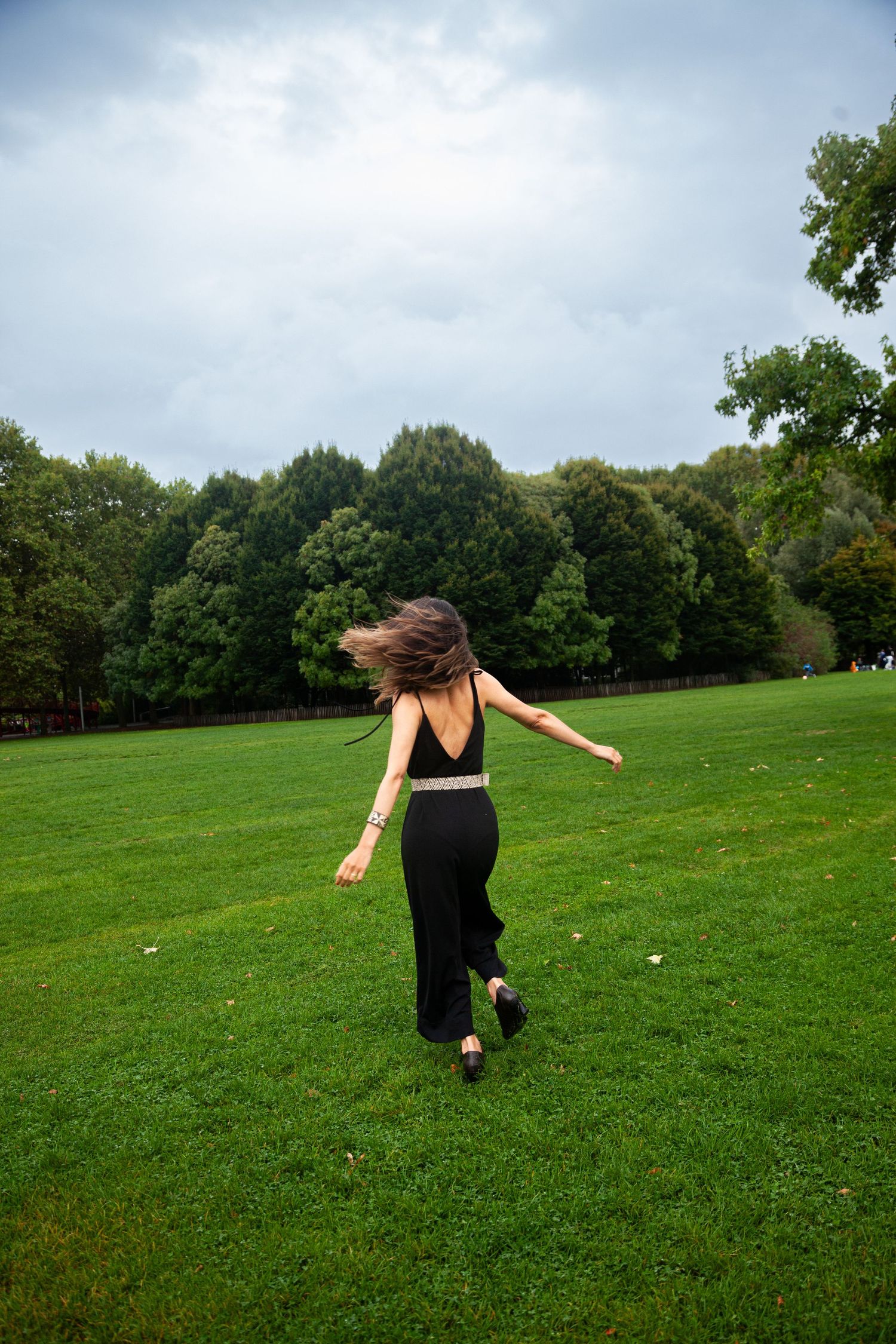 A person in a black outfit dances joyfully on a green lawn surrounded by trees.