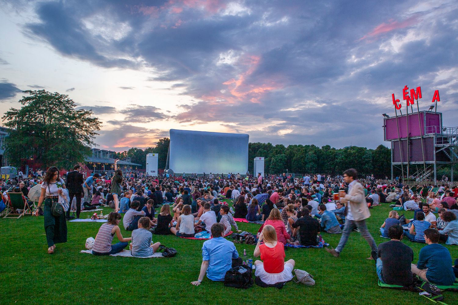 Crowd gathered on lawn at dusk for outdoor movie screening with pink-lit tower in background.