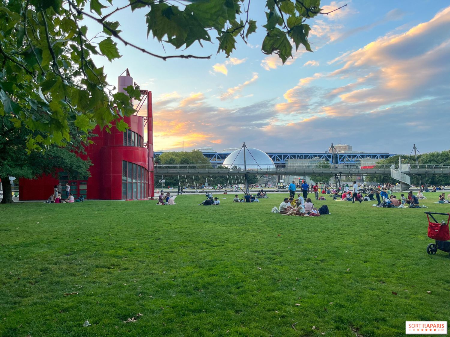 Red building and glass dome structure against sunset sky with people lounging on grass.