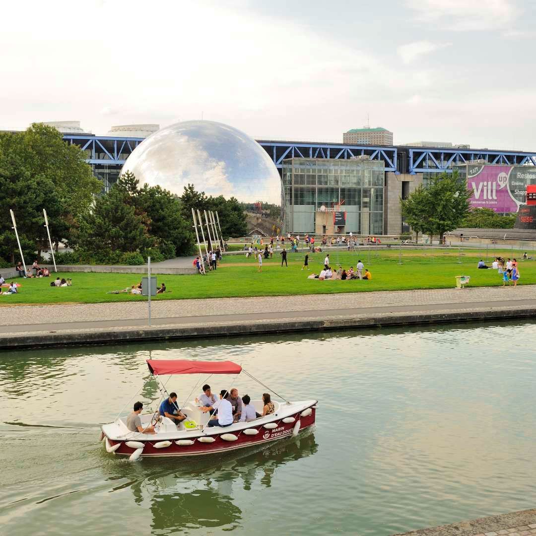 A large geodesic dome structure overlooks a waterfront where people relax and boat on a summer day.