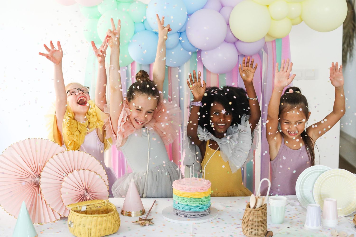 Group enjoying colorful birthday celebration with rainbow cake and pastel balloon decorations.