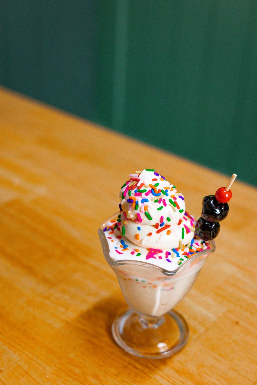 A milkshake topped with whipped cream, colorful sprinkles and a maraschino cherry sits in a glass on a wooden table.