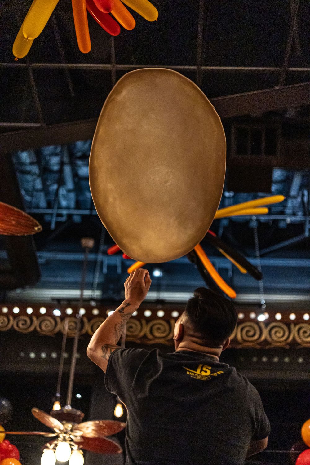 A staff member balances a large pizza dough in the air at a restaurant with decorative lighting.