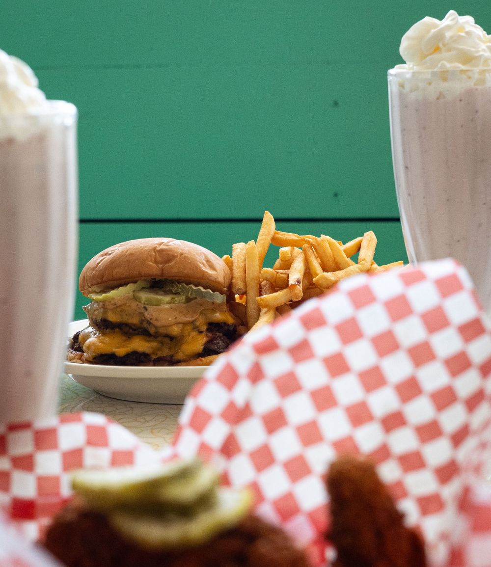 Cheeseburger with french fries and two milkshakes served on a green background with red checkered paper.