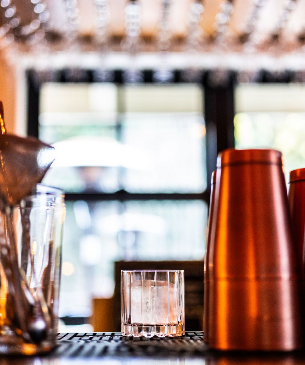Cocktail shaker and glass on a bar counter with a blurred window background.