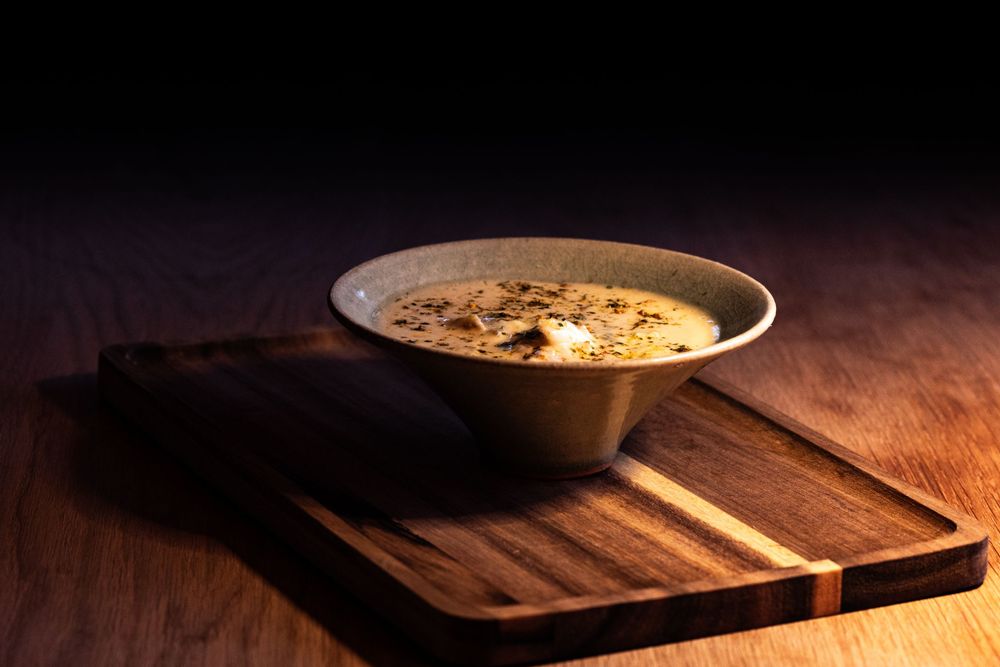 Bowl of creamy soup garnished with herbs and spices on rustic wooden serving board under dramatic lighting.