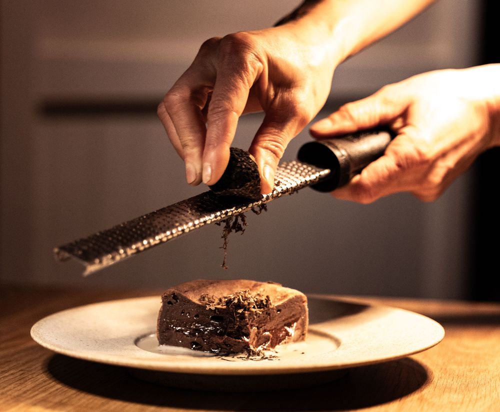 Hands grating chocolate over a dark chocolate dessert on a white plate in warm lighting.