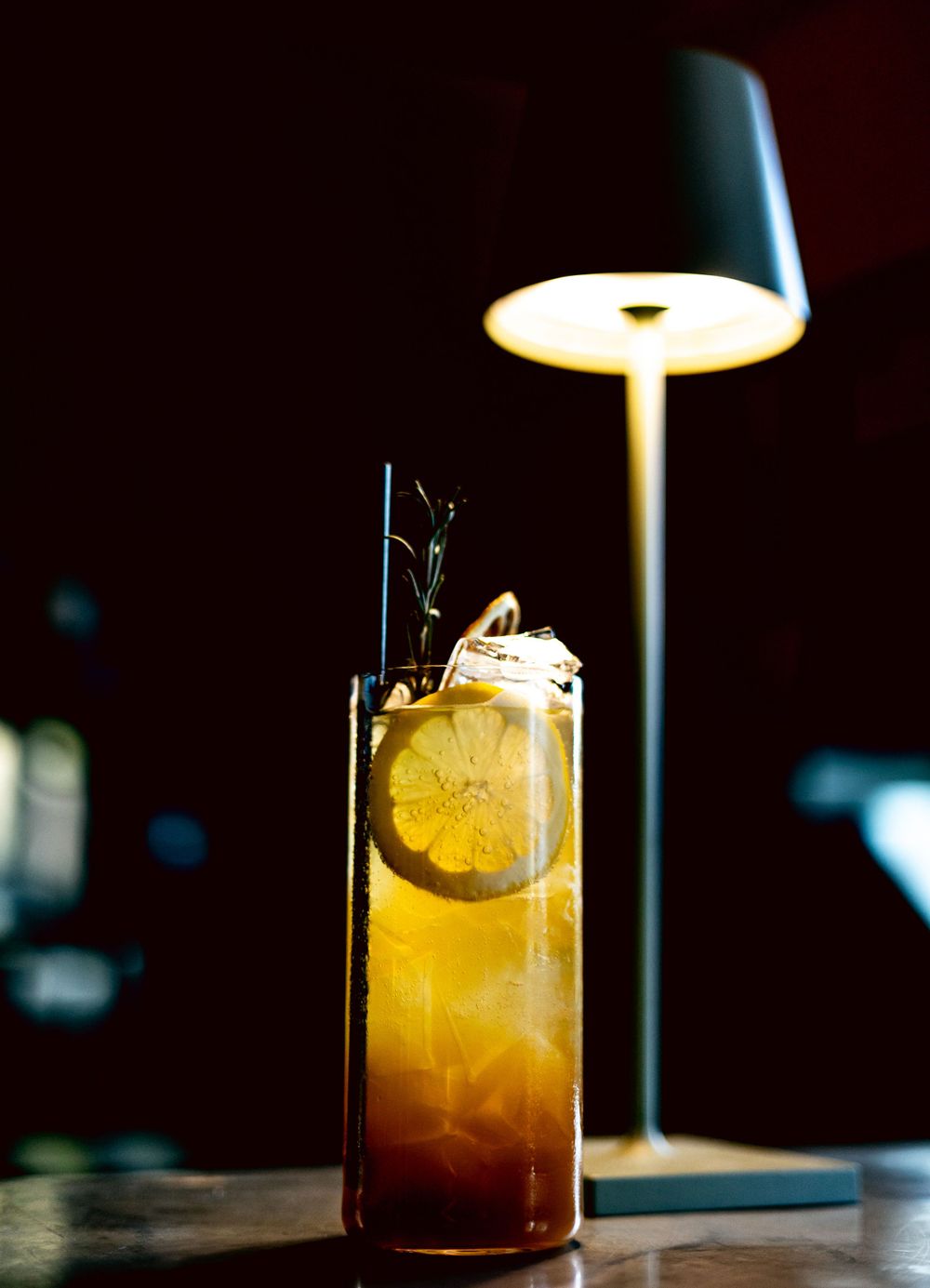 Iced cocktail with lemon garnish and herbs served in a tall glass under a modern pendant lamp.
