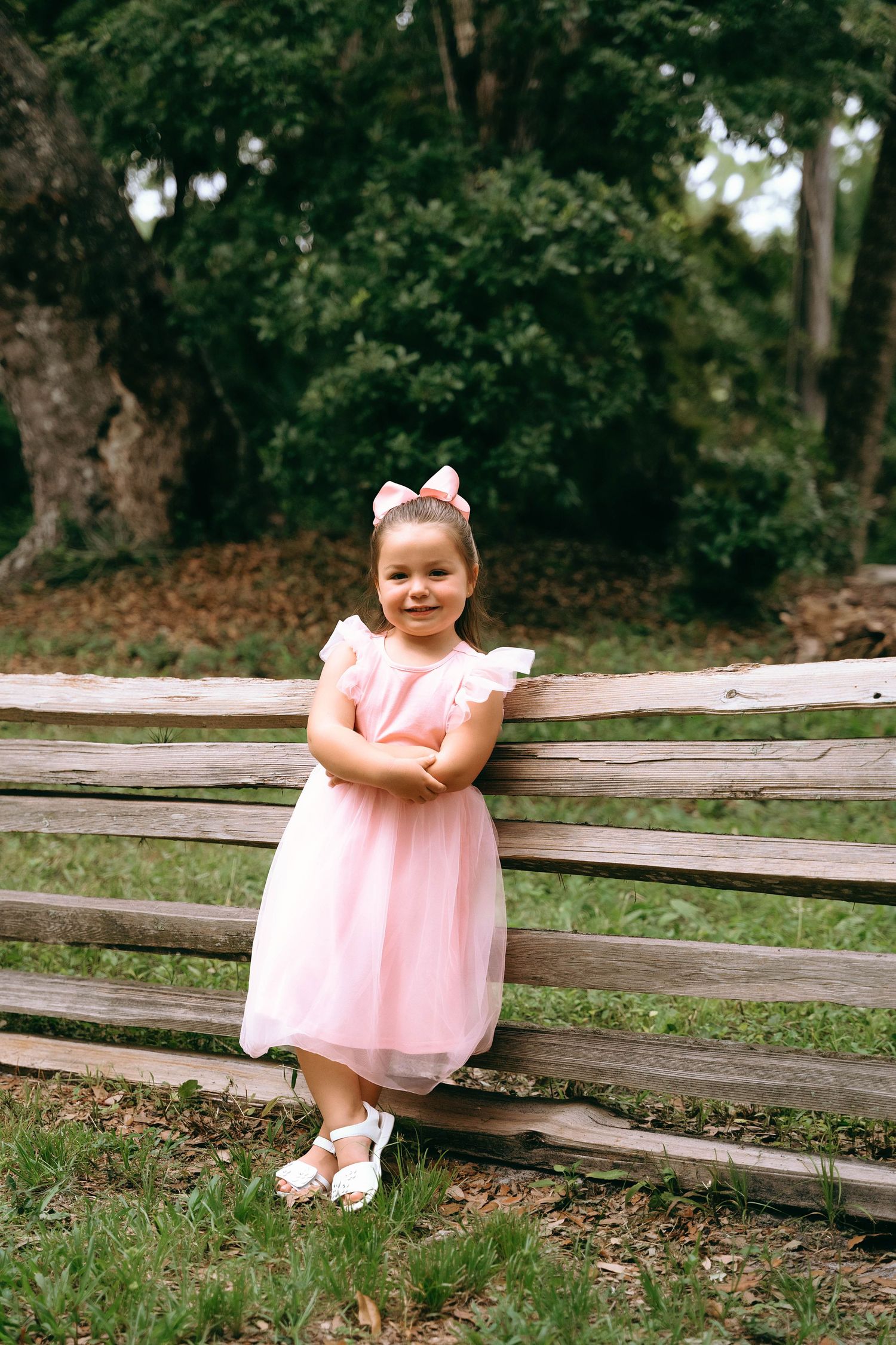 A child in a pink dress sits on a rustic wooden fence surrounded by lush green trees and grass.