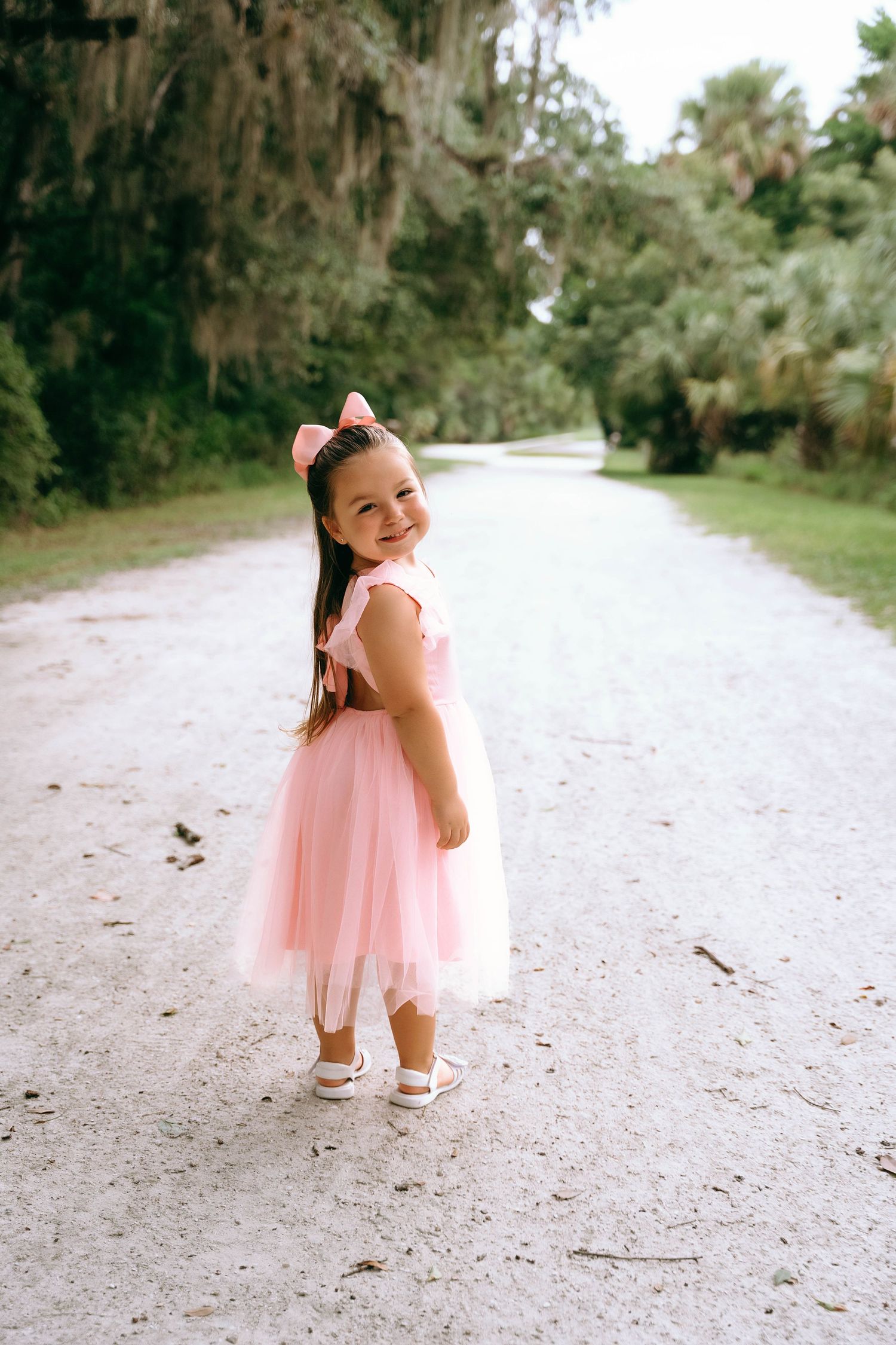 A young dancer in a pink tulle dress twirls playfully on a sandy path lined with Spanish moss and palm trees.