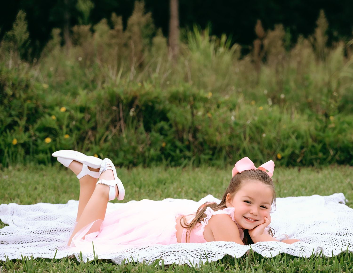 Child lying on a white blanket in the grass wearing a pink outfit and white shoes during a summer day.