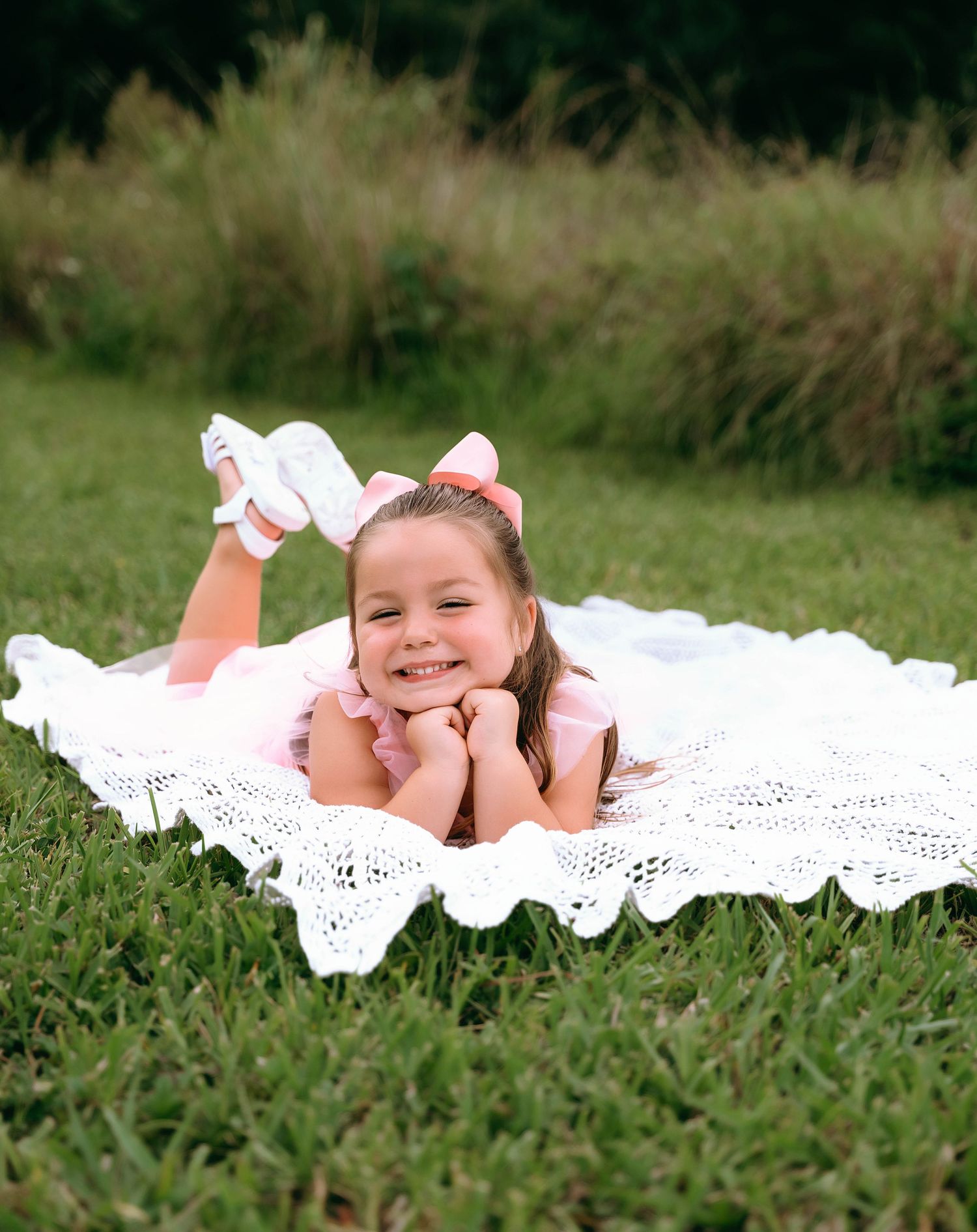 A baby lays on a white crocheted blanket in the grass while smiling and giggling during an outdoor photo session.
