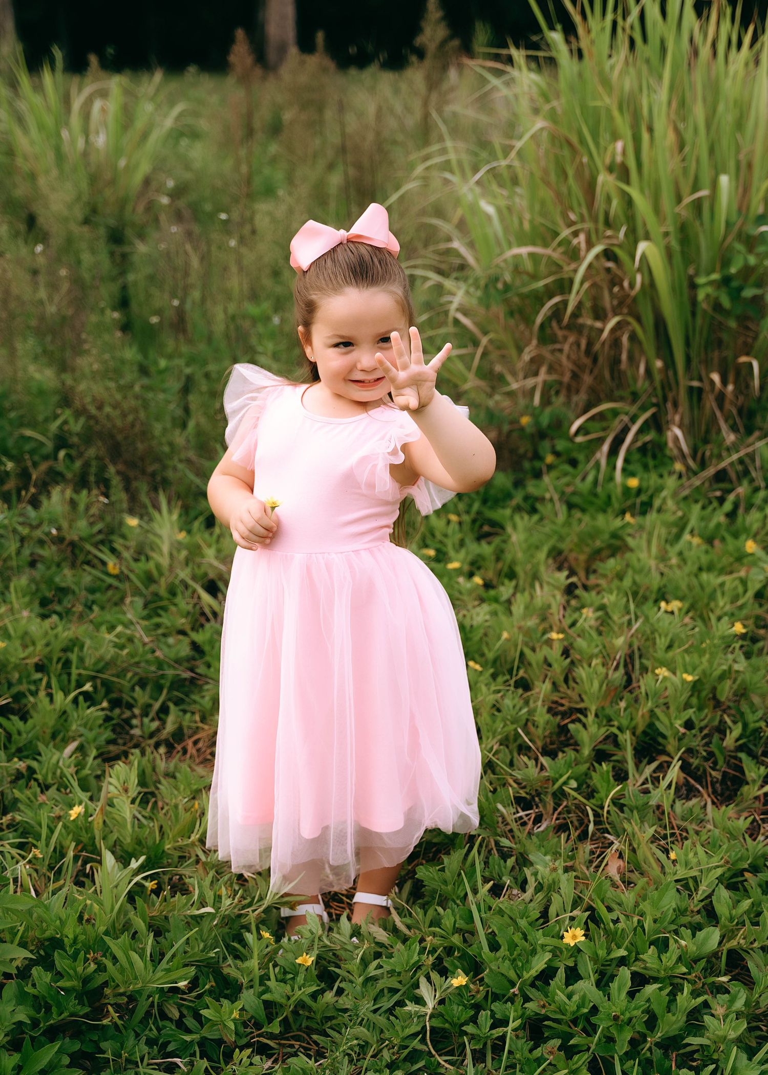 A small figure in a pink tulle dress and bow poses on green grass surrounded by tall ornamental grasses.