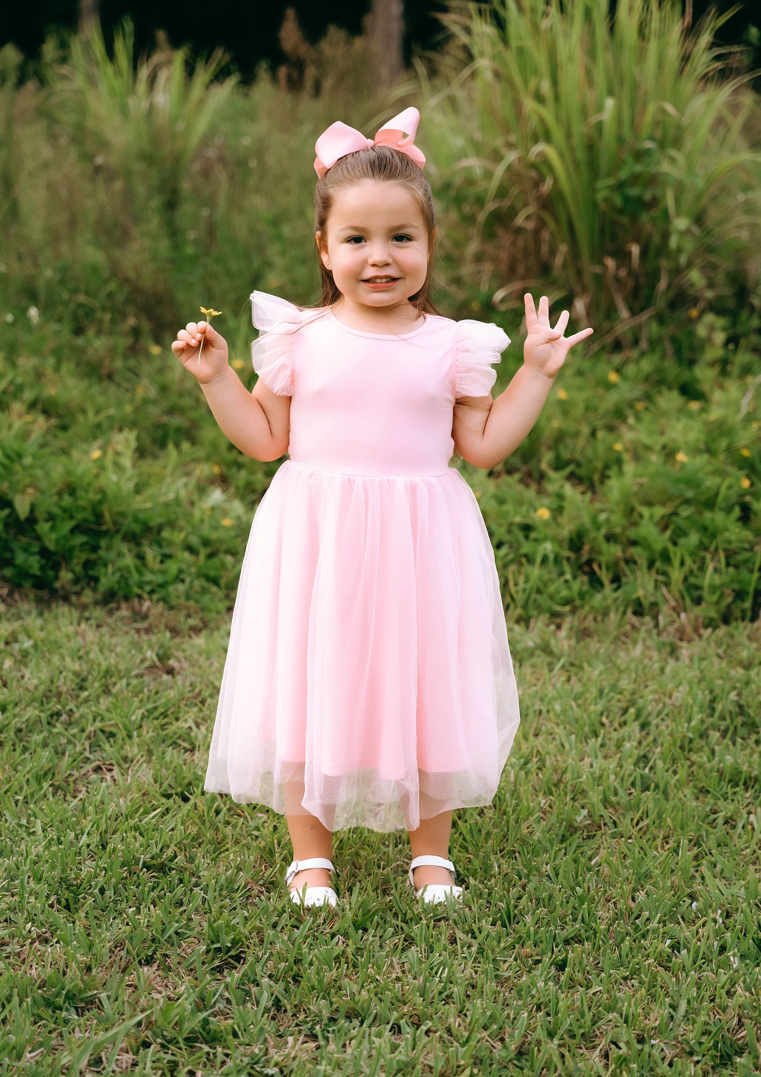 A small child wearing a pink tulle dress and white hair bow stands on grass with hands raised joyfully.