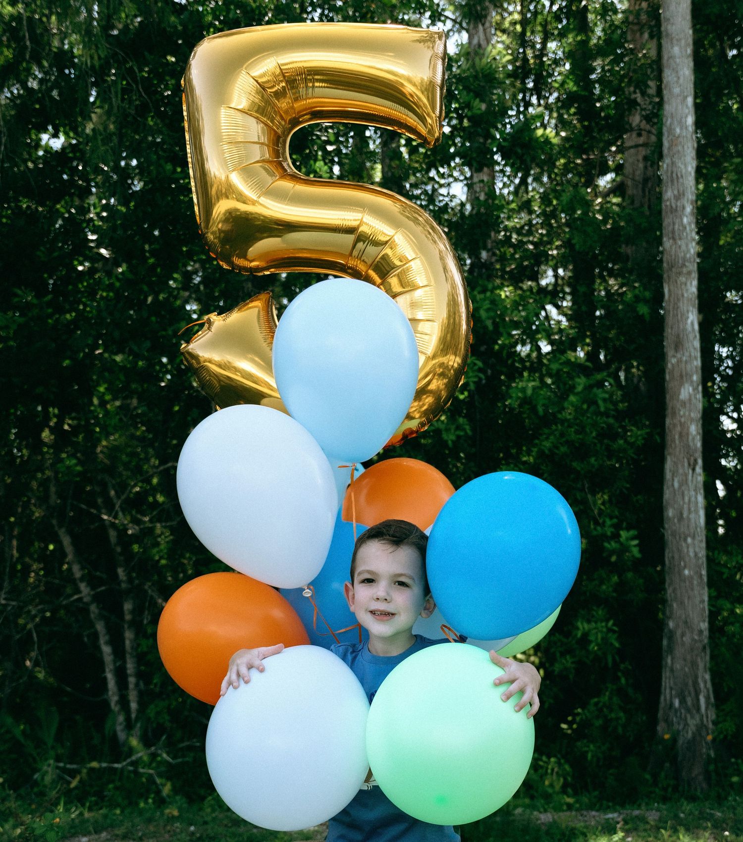 Colorful party balloons including gold number balloons and blue, white and orange balloons arranged in woodsy setting.