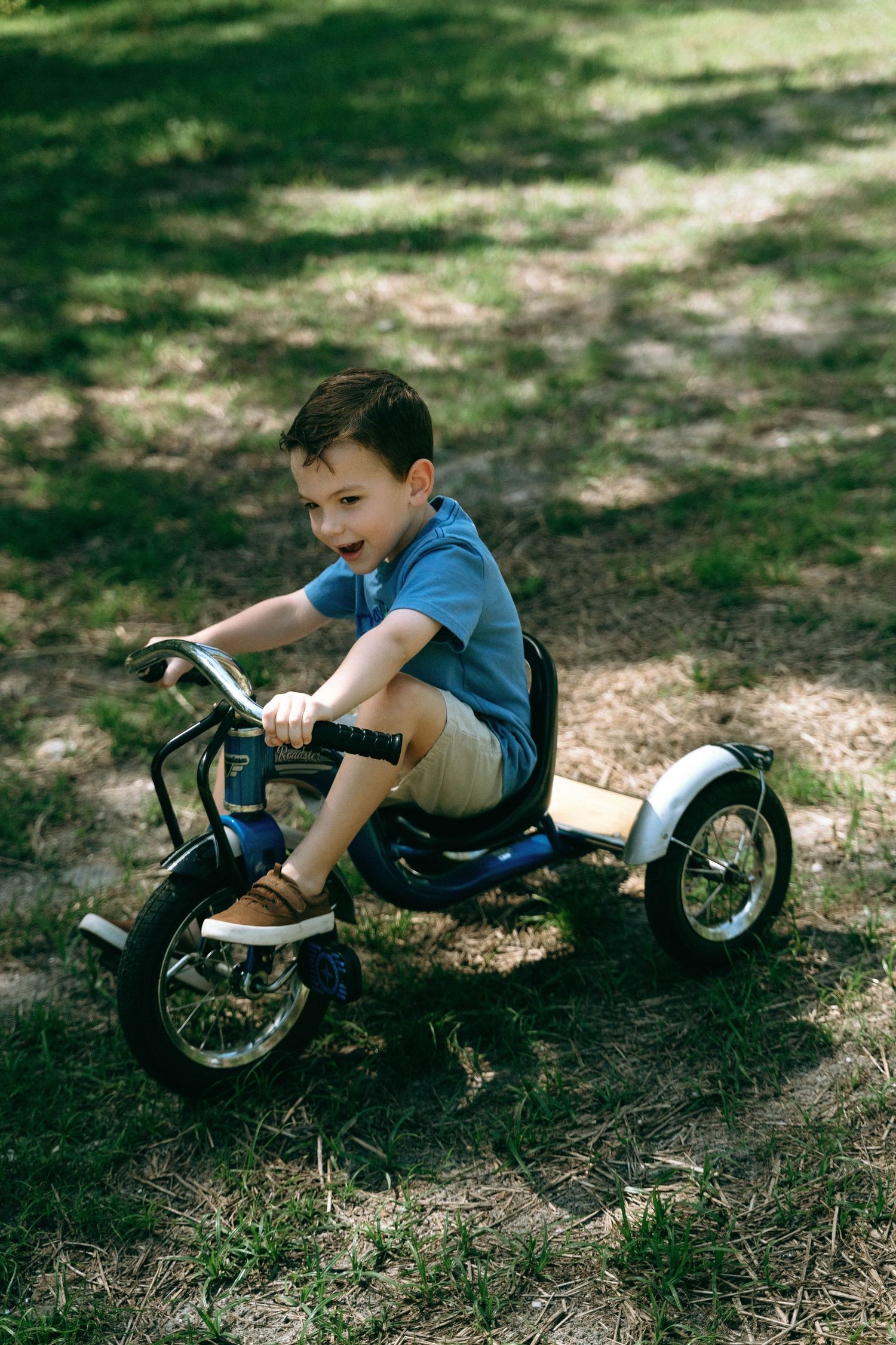 A child rides a vintage-style silver tricycle on a shaded outdoor path.