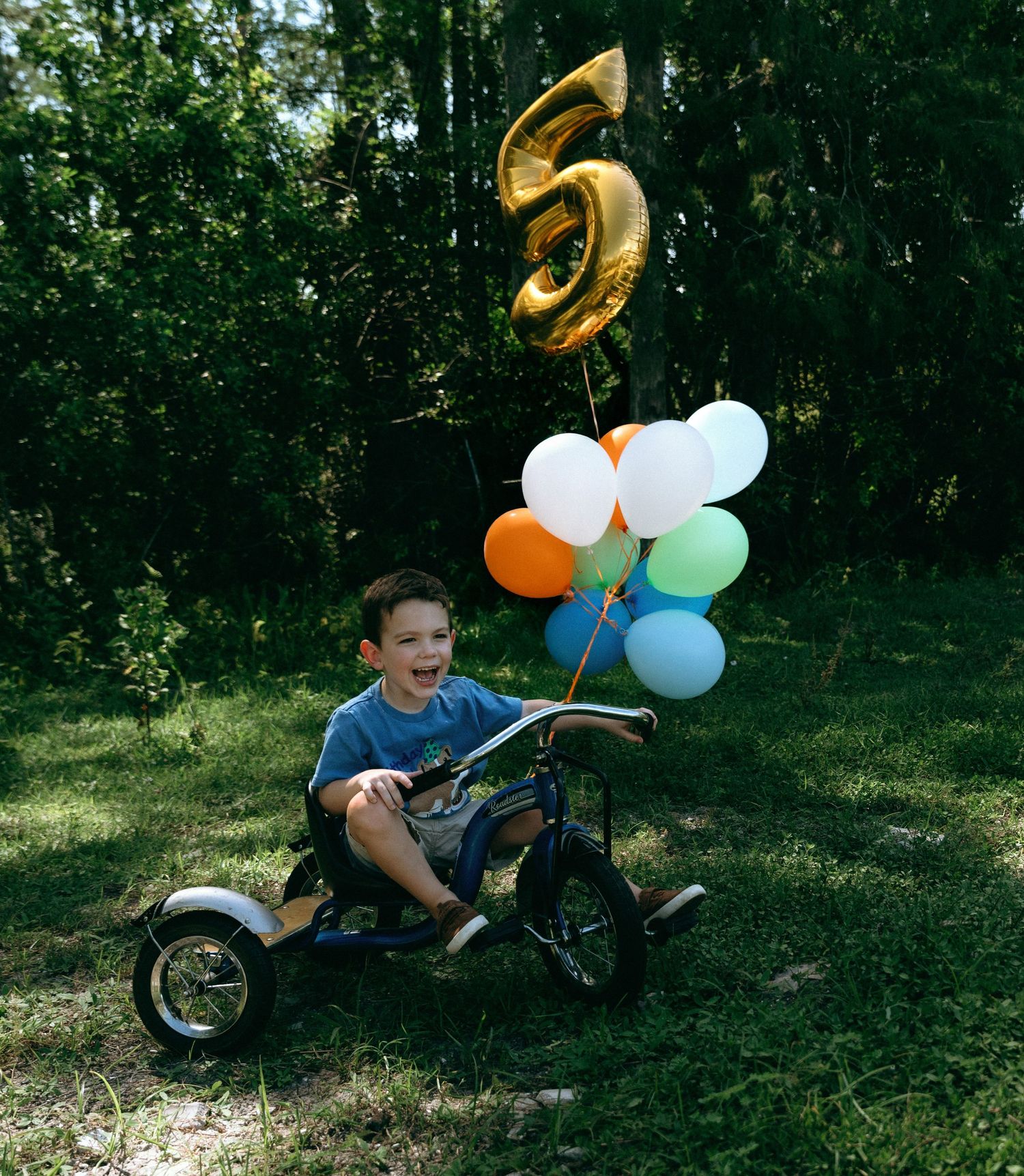 A child rides a tricycle decorated with colorful balloons and a gold number 5 balloon in a leafy outdoor setting.
