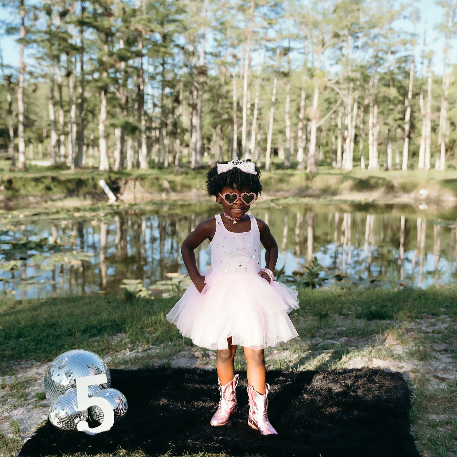 A joyful celebration next to a pond with sparkly boots, a tutu dress, and 3-shaped balloon in a wooded setting.