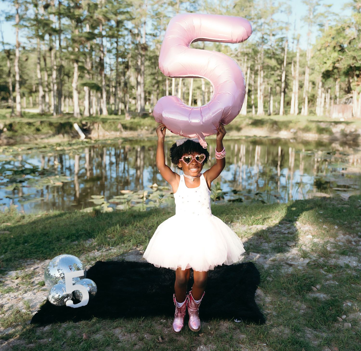 A festive birthday moment outdoors with a pink number 5 balloon and white tutu dress by a serene pond.