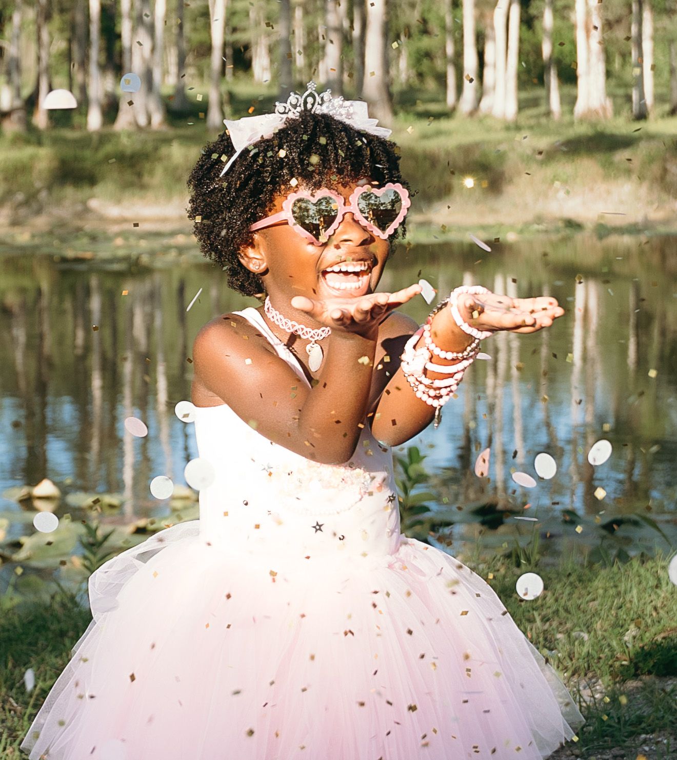 A person in a fluffy white dress and sunglasses sits on grass near a pond while enjoying a sunny afternoon.