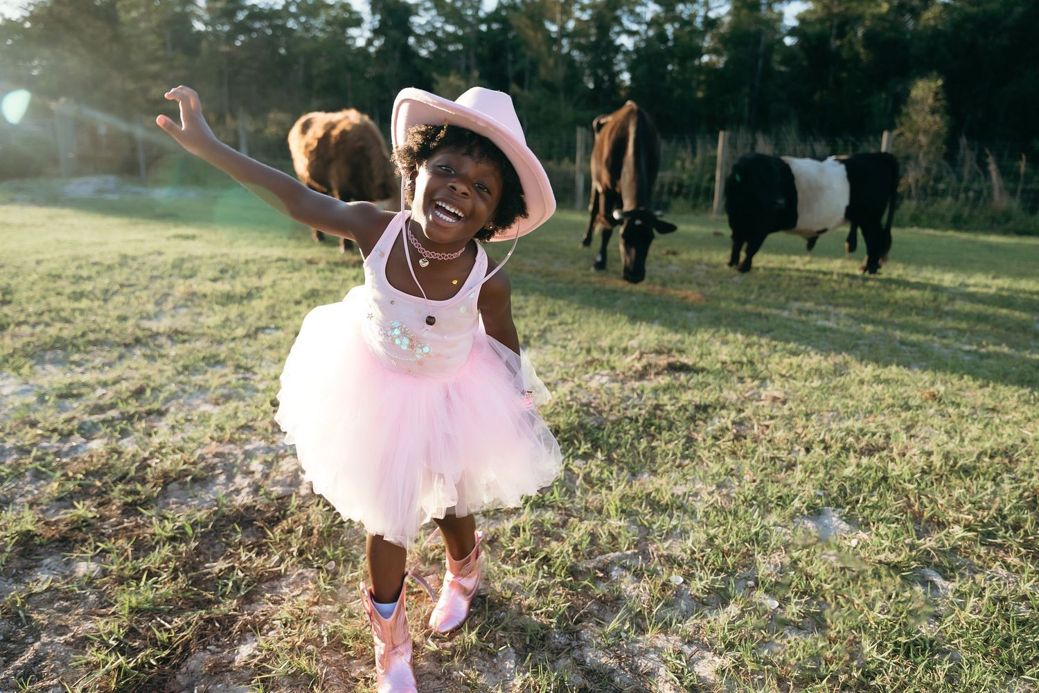 A happy person in a pink tutu and cowboy boots dances in a field with horses grazing in the background.