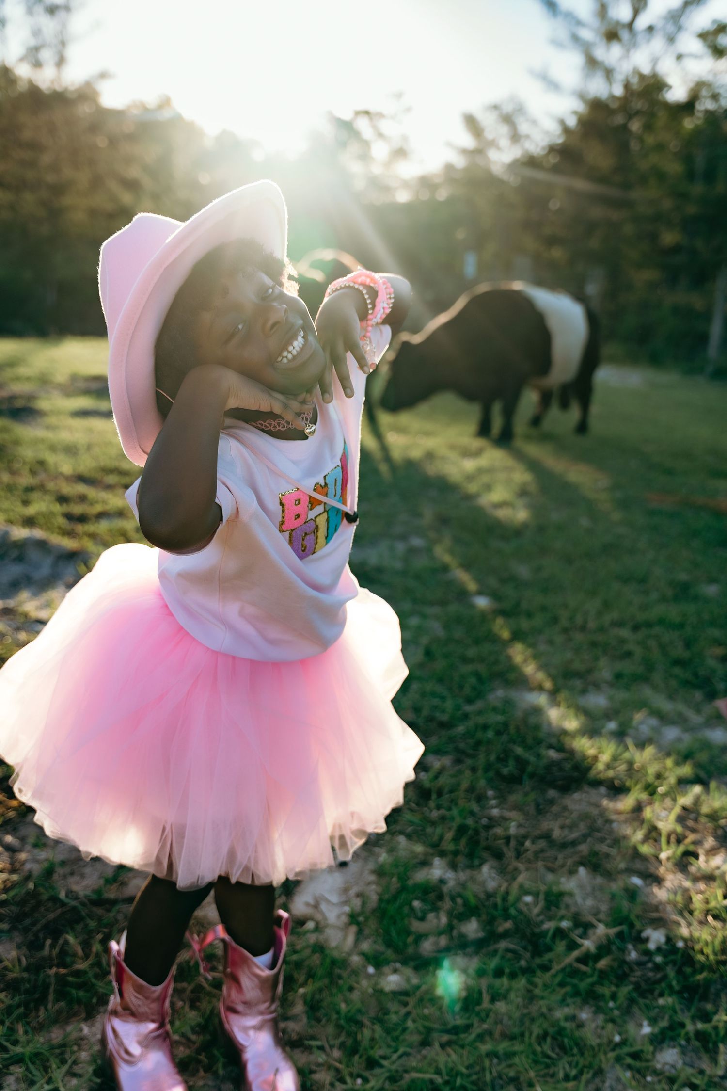 Child in pink tutu and cowgirl boots enjoys a sunny day in a field with horses grazing in the background.
