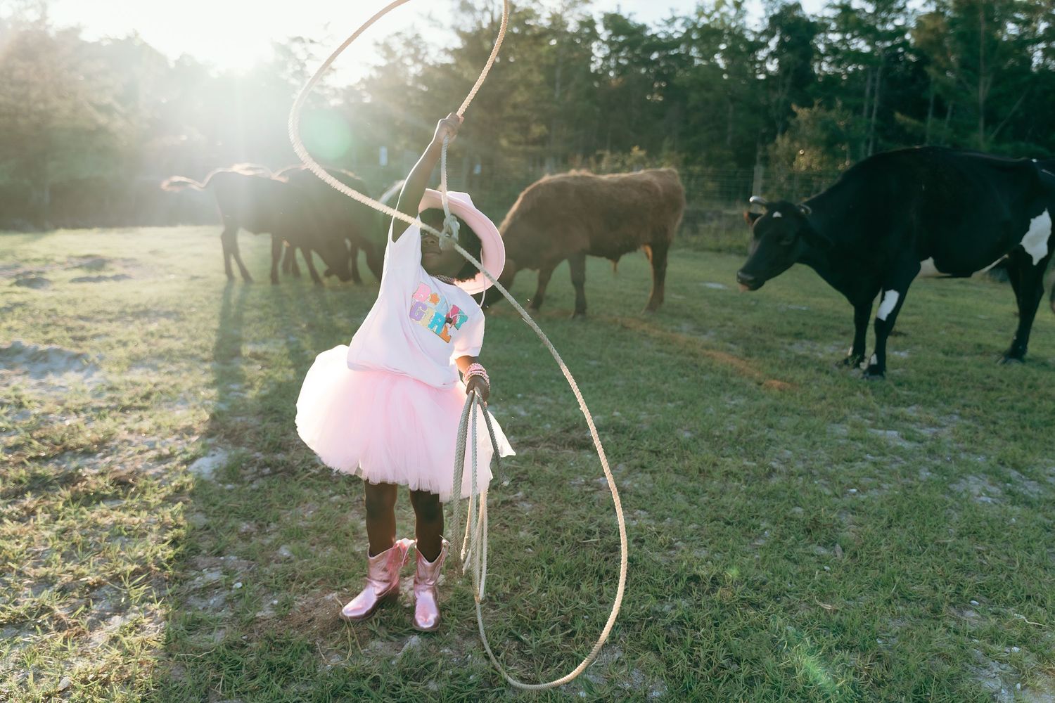 Child in pink tutu carrying cowboy rope stands in field with grazing cattle at golden sunset.
